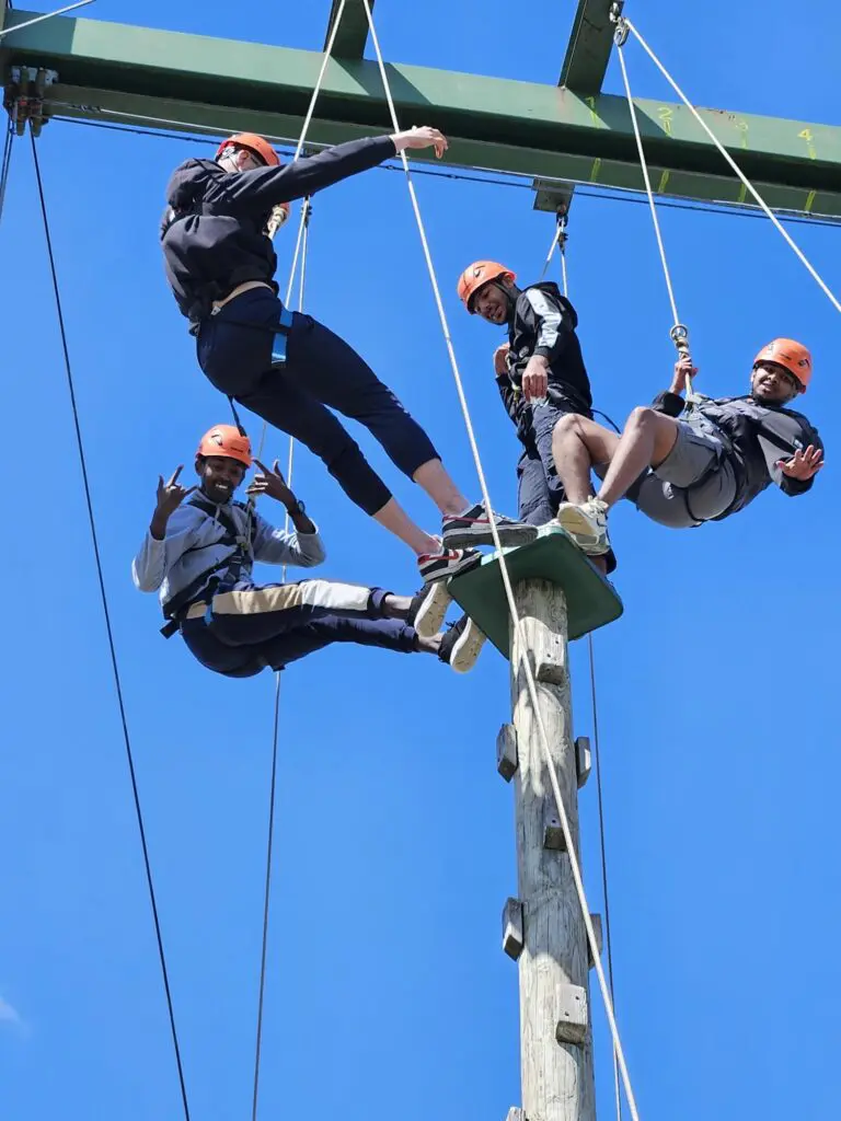 4 of our Gaisce Bronze participants balancing on a high stand in helmets and harnesses on abseiling ropes. There is a blue sky in the background.