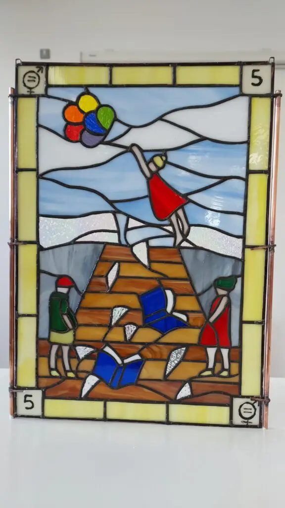 Photograph of a stained glass piece depicting SDG 5 - Gender Equality. The work shows a staircase with open books falling down it, two women stand at the bottom of the stairs whilst at the top a woman reaches for a bunch of balloons that are flying in the sky.