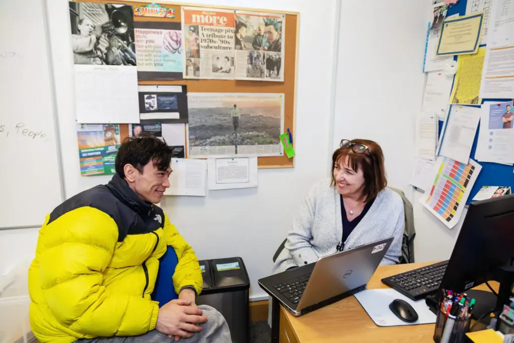 A Community Training Centre participant and a tutor sit at a desk with a laptop on smiling at each other. Behind them the classroom walls are decorated in newspaper clippings and posters. Photographed by Paul Reardon.