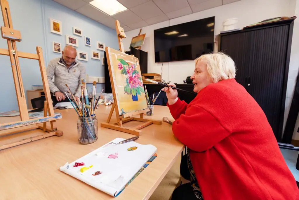 Participants from our Adult Ed art class prepare for class. One participant is painting a still life of a blue vase full of pink flowers and green leaves. Photographed by Paul Reardon.