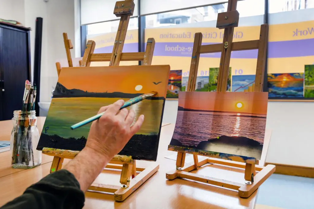 A hand holding a paint brush, painting a sunset over a bay from an inspiration photo. The painting sits on an easel on a table, with other paintings shown in the background. Photographed by Paul Reardon.