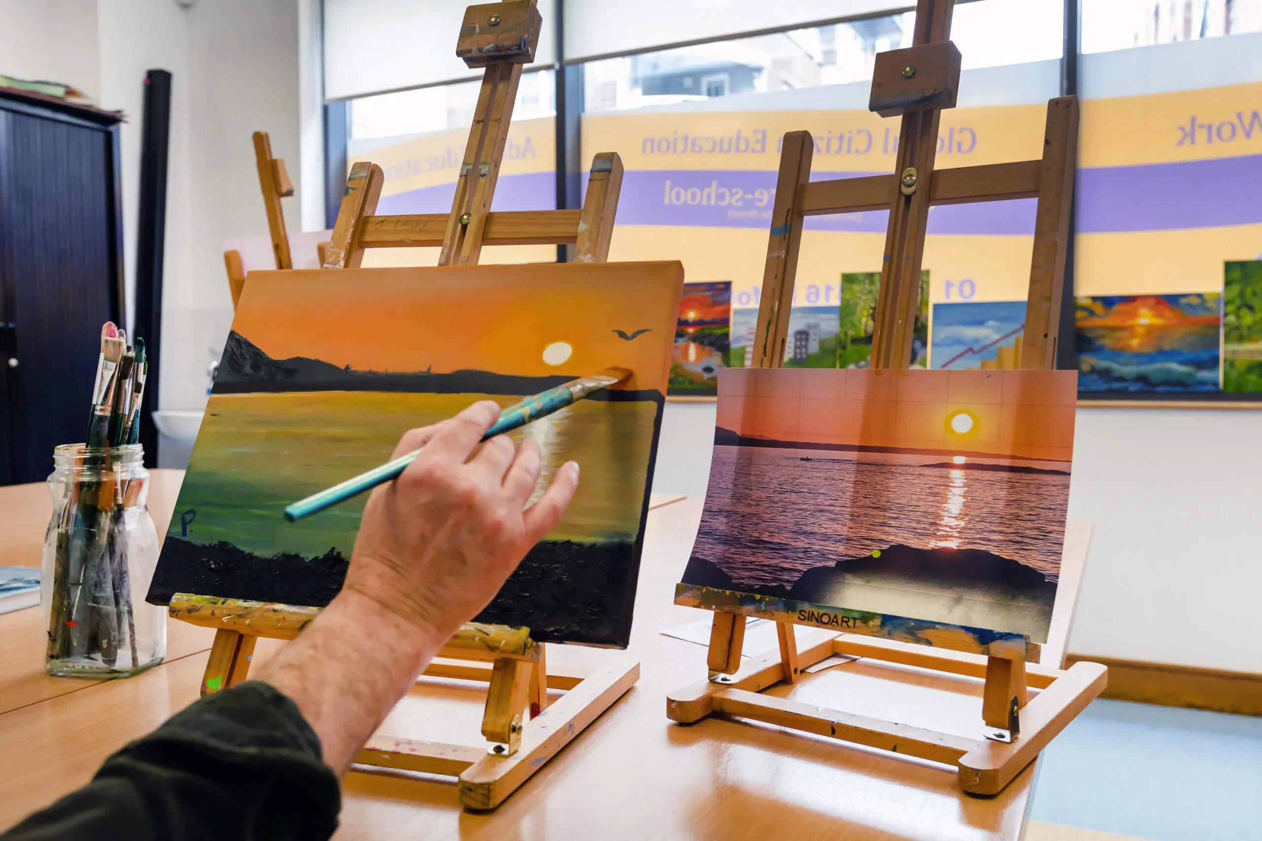 A hand holding a paint brush, painting a sunset over a bay from an inspiration photo. The painting sits on an easel on a table, with other paintings shown in the background. Photographed by Paul Reardon.