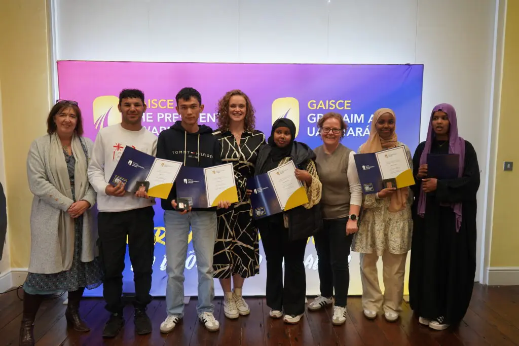 Our Gaisce participants receive their award, smiling at the camera whilst holding up their certificates and their bronze awards. Pictured with two LYCS staff members in front of a purple and blue Gaisce poster with gold lettering.