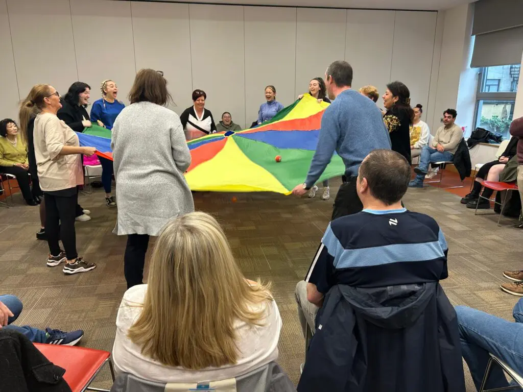 A staff training activity where participants must keep a number of balls rolling on a colourful parachute they're holding. A number of particpants sit in an outer circle and watch in amusement.