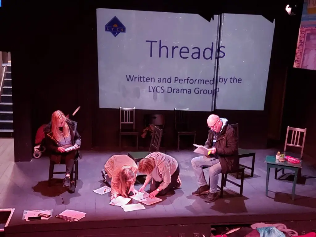 Our Theatre of the Oppressed group sit and crouch on stage surrounded by chairs and scripts. Behind them is a screen that reads 'Threads: Written and Performed by the LYCS Drama Group'.
